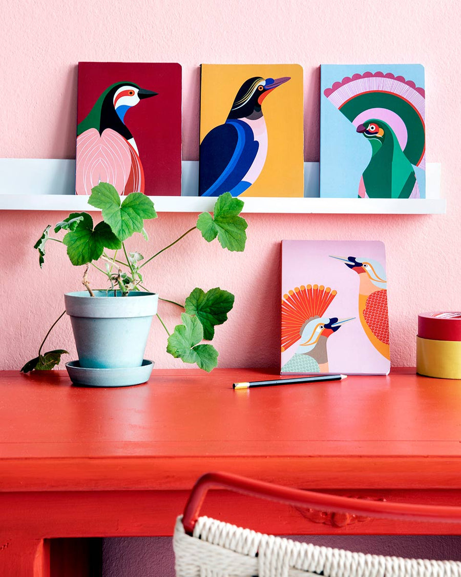 Studio Roof eco-friendly Paradise Bird Notebooks lined up on a book shelf above a red work desk