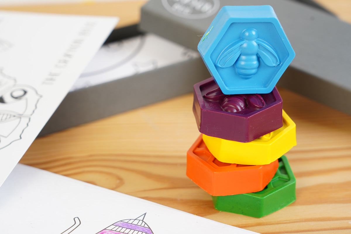 Close up of the hexagonal crayon bug bee crayons set stacked in a tower on a wooden background next to their grey box