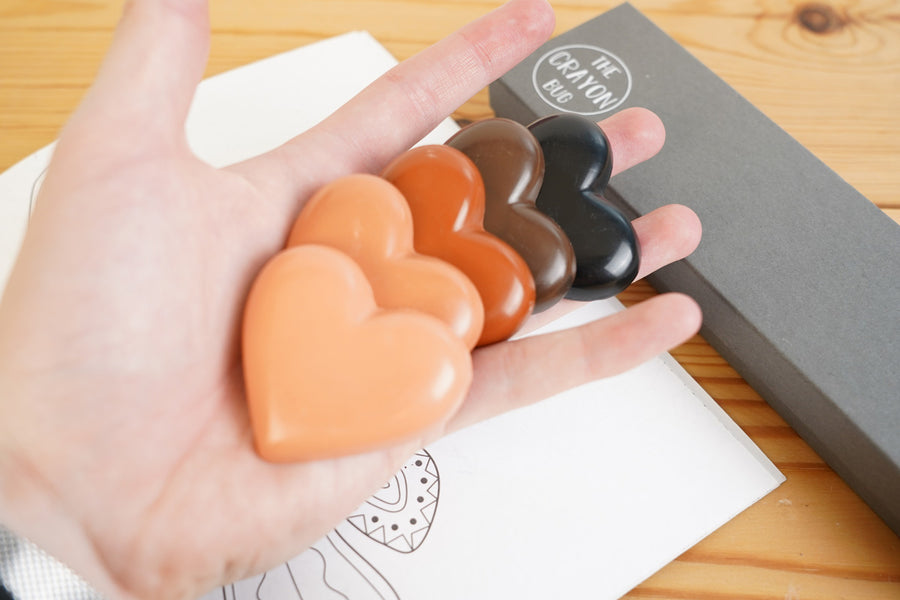 Close up of a hand holding the Crayon Bug skin tone heart crayons over a wooden background and colouring book