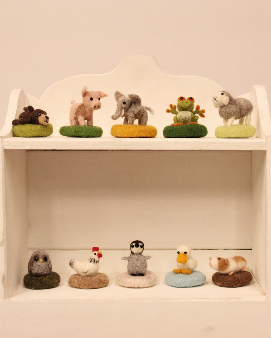 The Makerss - Amiguwoolli Tiny Mini Needle Felt Figures. 10 mini needle felt figures lined in two rows. Sat on top of a wooden box is the mini pig