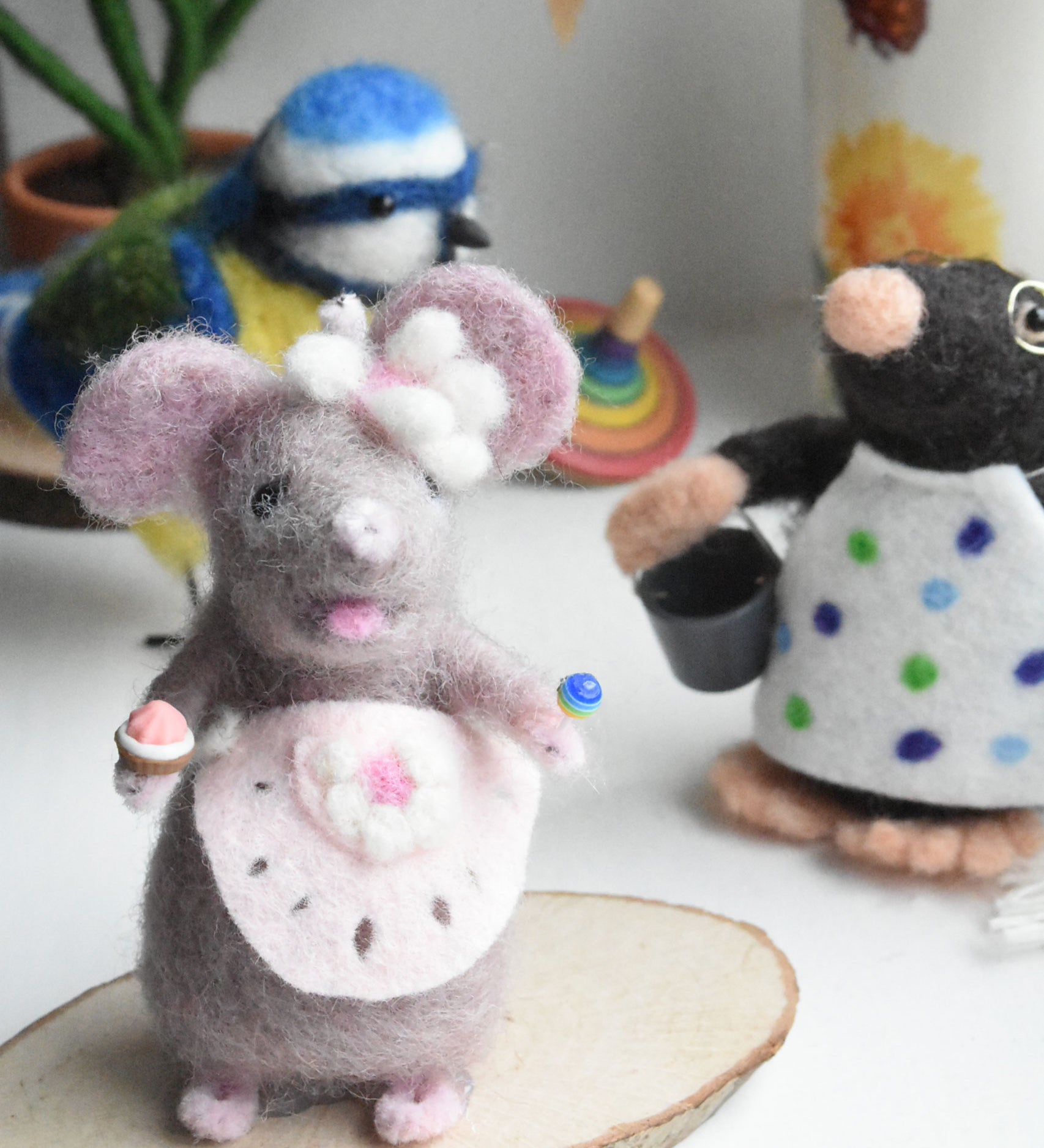 A felted mouse figure wearing a pink apron and holding a cake and lollypop in it's hands. The figure has been created using The Makerss Candy Mouse Needle Felt Craft Kit.