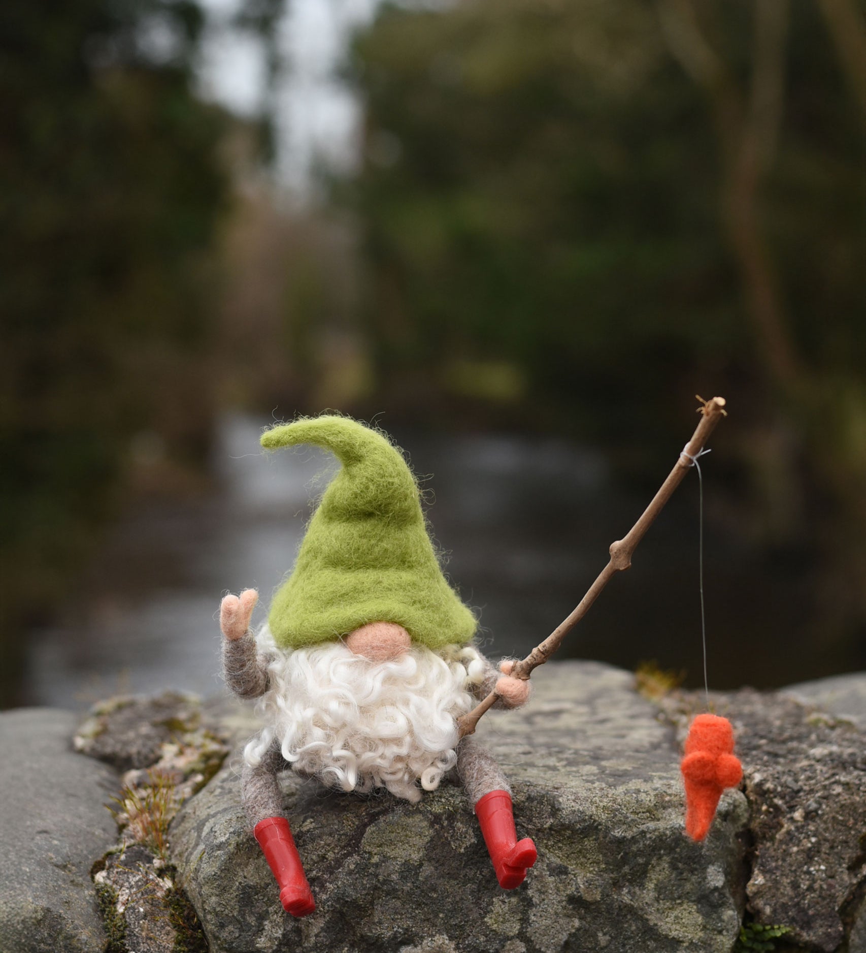 A felted gnome in a seated position with a fishing rod fashioned from a wooden stick created using The Makerss - Small Fishing Gnome Needle Felt Kit. 