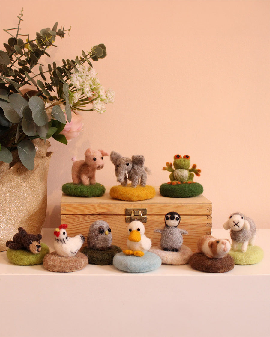 The Makerss - Amiguwoolli Tiny Mini Needle Felt Figures. 10 mini needle felt figures lined in two rows. Sat on top of a wooden box is the mini pig