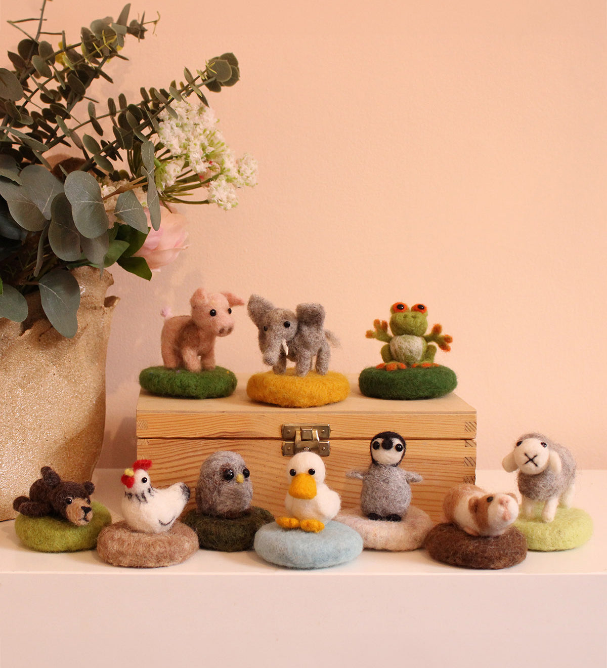 The Makerss - Amiguwoolli Tiny Mini Needle Felt Figures. 10 mini needle felt figures lined in two rows. Sat on top of a wooden box is the mini pig