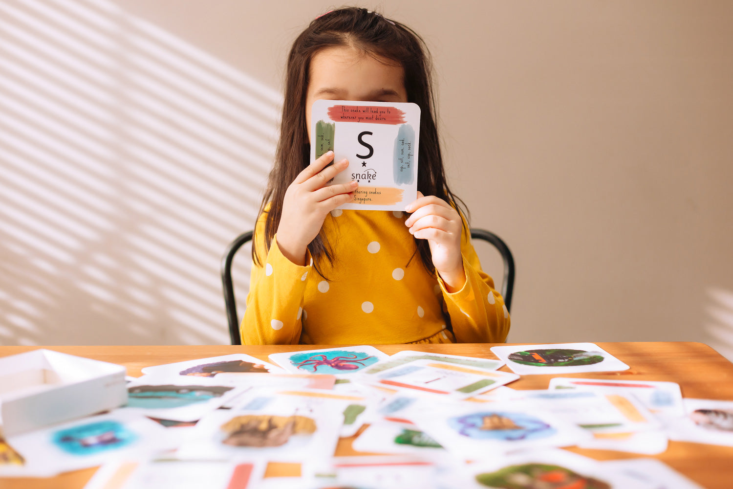 Child holding up a card with the letter S for snake on from the The Phive Enchanting Learning Phonics & Storytelling Cards - Set 1 with all the other cards laid out on the table in front of them