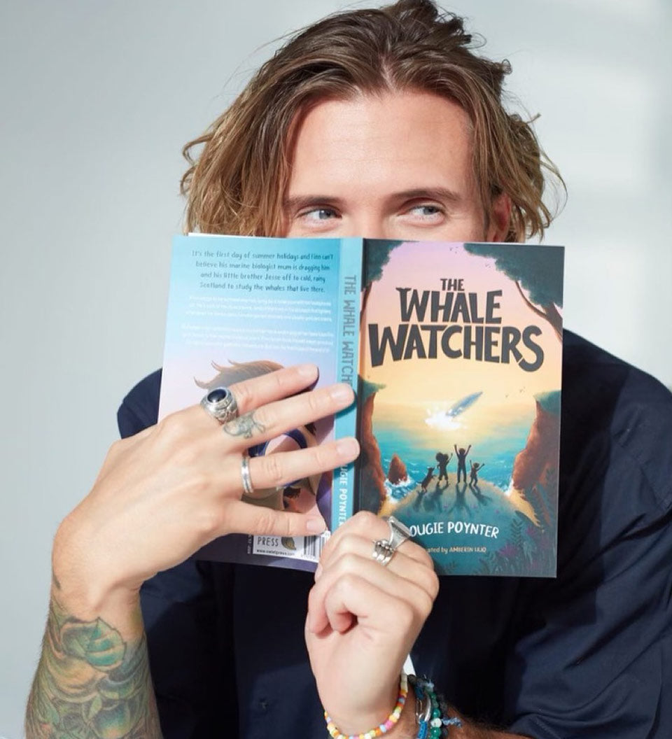 Author Dougie Poynter holding The Whale Watchers childrens book up in front of his face on a grey background