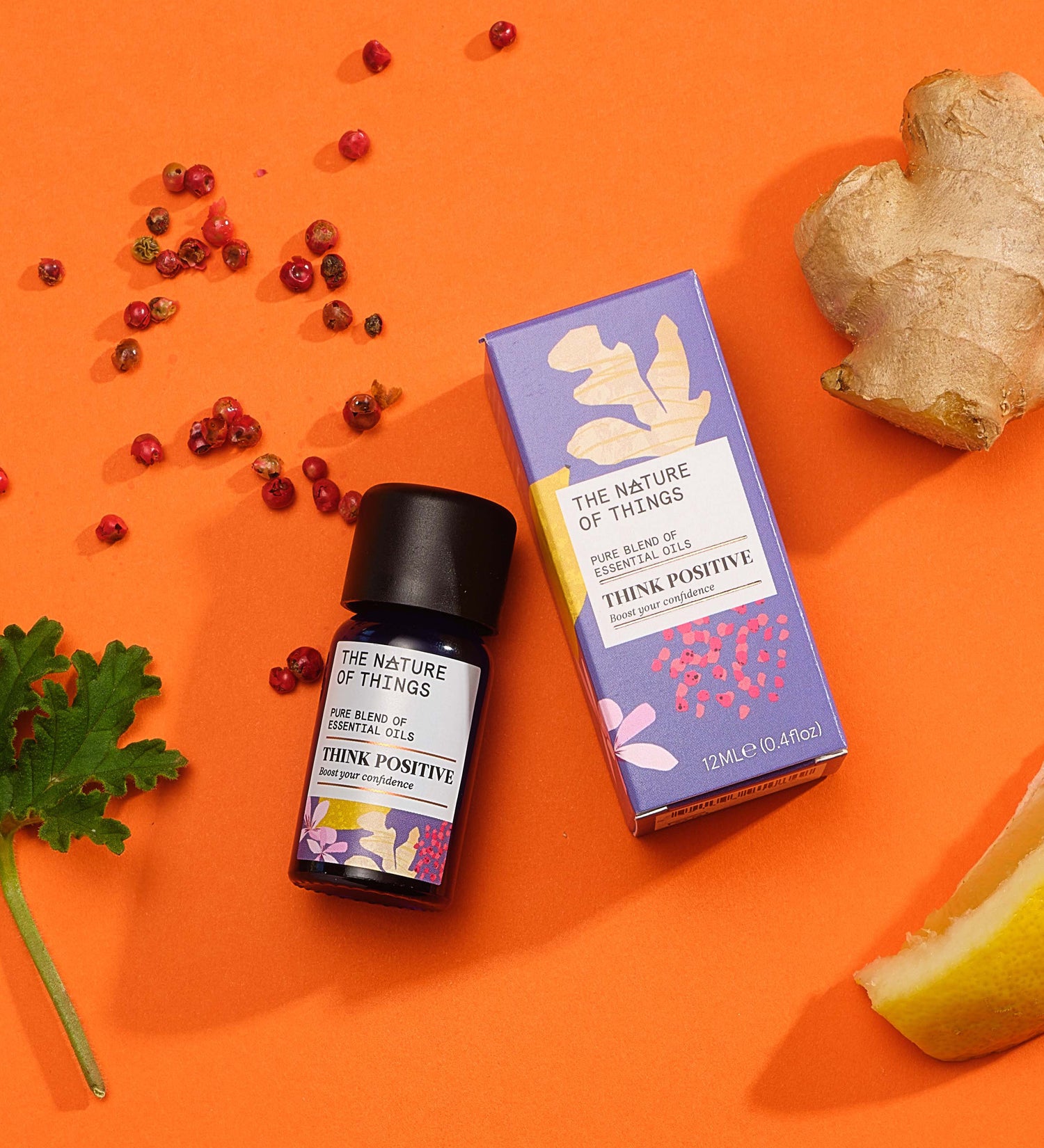 Think Positive pure essential oil blend in a blue glass bottle next to a decorative purple cardboard box, with peppercorns, ginger, lemon and geranium on an orange background. 