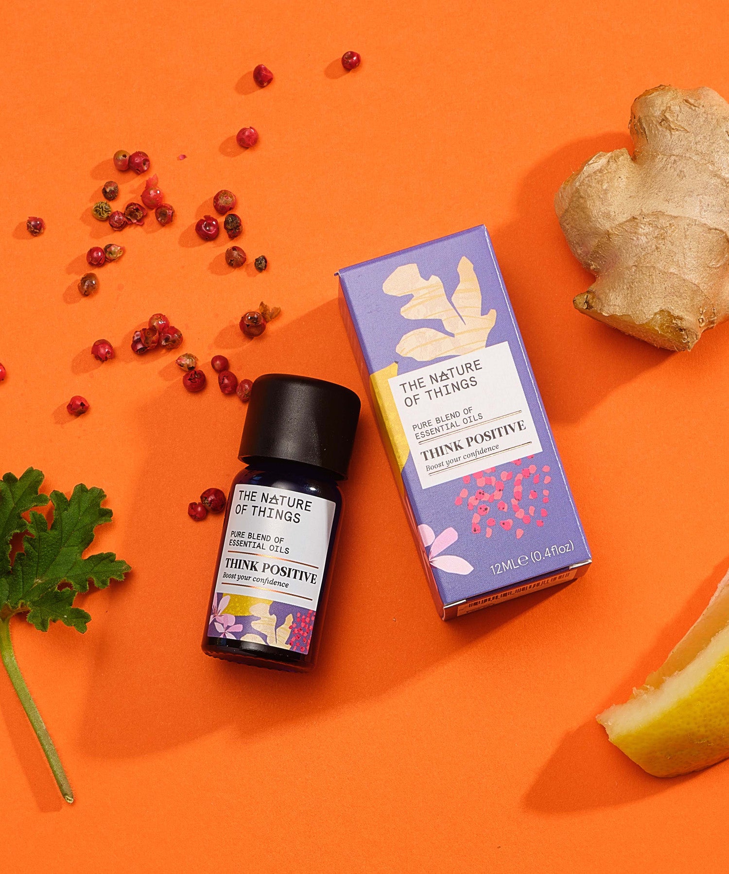 Think Positive pure essential oil blend in a blue glass bottle next to a decorative purple cardboard box, with peppercorns, ginger, lemon and geranium on an orange background. 