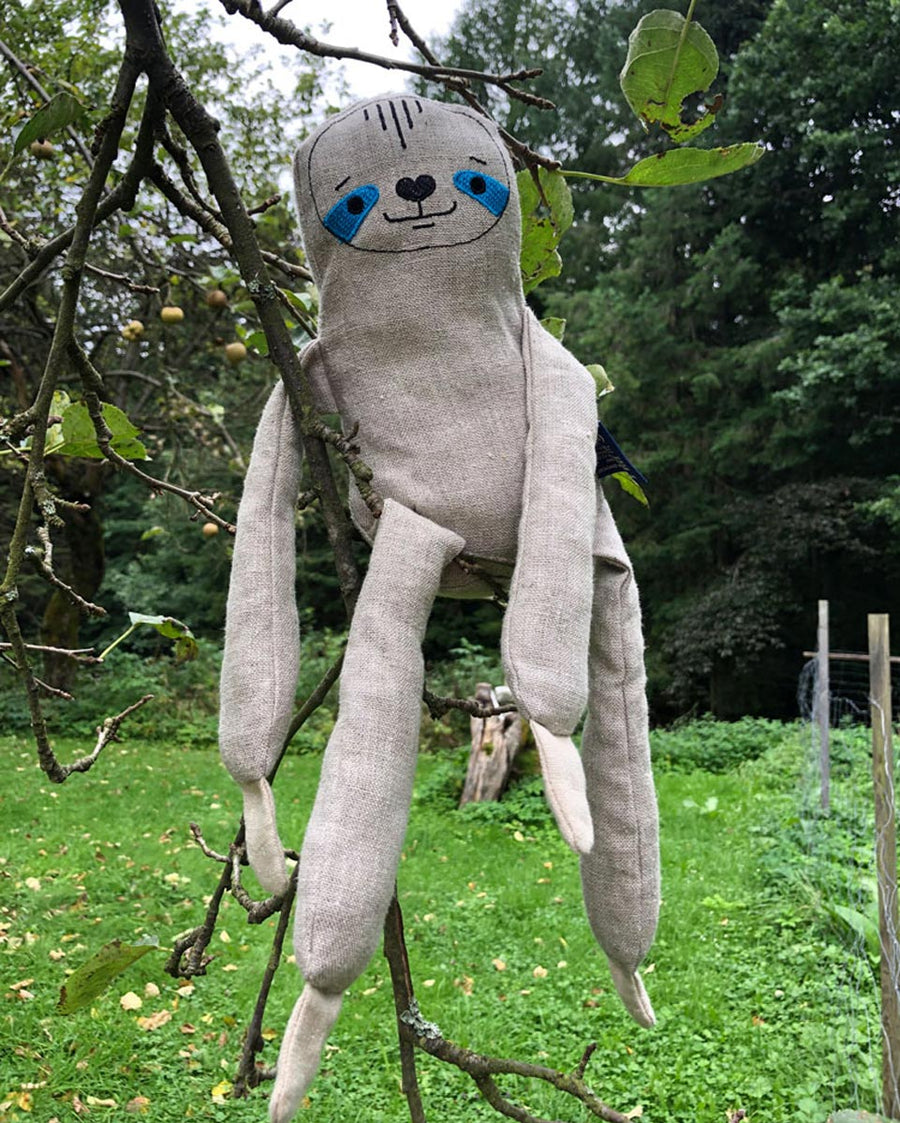 Tobe children's plush hemp sloth figure sitting on a tree branch in front of a field