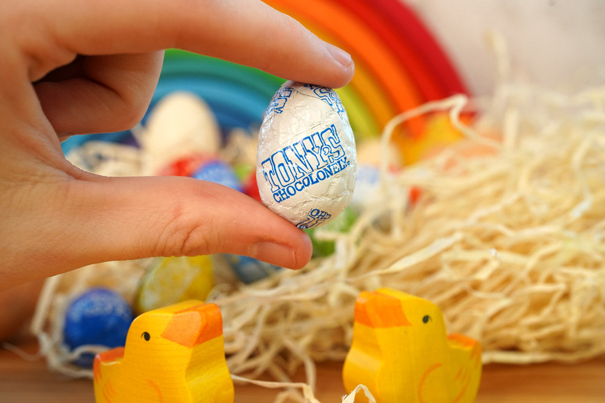 Close up of a hand holding a Tonys chocolonely fairtrade mini easter egg in front of some wooden chick toys