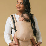 Woman stood on a beige background with a baby in a Tula forward facing baby carrier