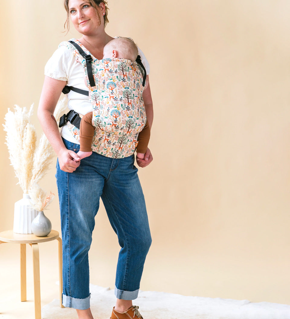 Woman stood in front of a beige wall carrying a baby in a Tula charmed free to grow baby carrier