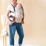 Woman stood in front of a beige wall carrying a baby in a Tula charmed free to grow baby carrier