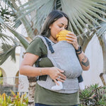 Close up of a woman kissing a young baby in the Tula eco-friendly soft free to grow baby carrier in front of a large palm tree