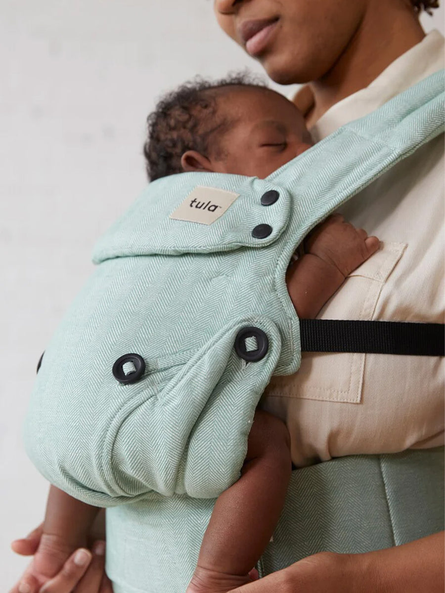 A baby is fast asleep in the Explore Eucalyptus Carrier. Parent is gently touching the babies feet and looking down at the baby