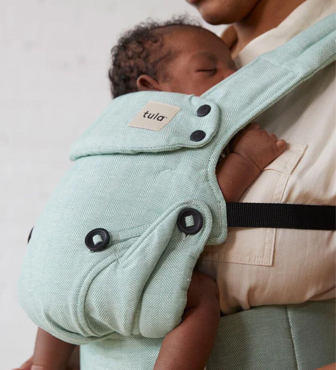 A baby is fast asleep in the Explore Eucalyptus Carrier. Parent is gently touching the babies feet and looking down at the baby