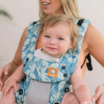 Tula - A woman carries a child on her chest wearing a Coast Paradise - Explore Baby Carrier. This is a front-on lifestyle shot with a soft beige background. The focus is on the button detail on the carrier.