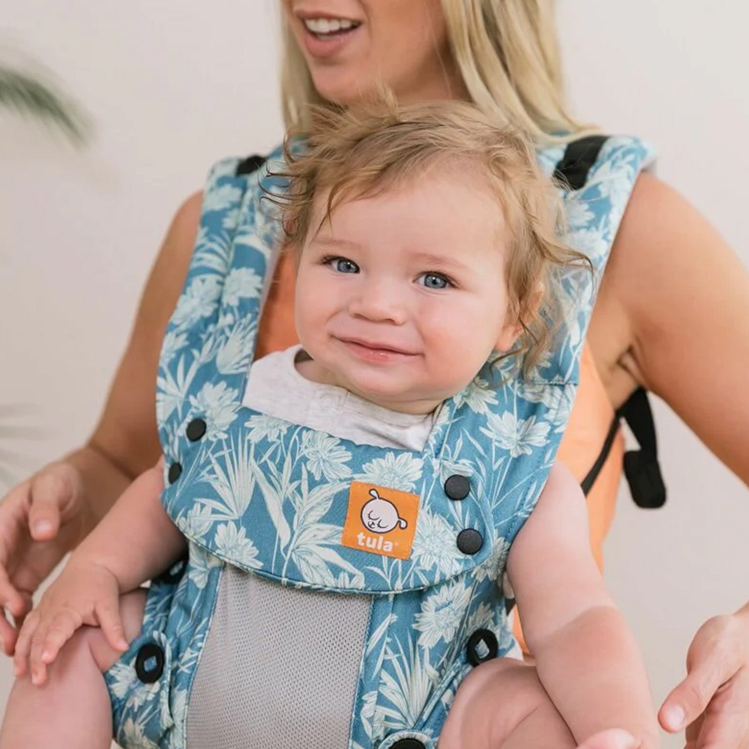 Tula - A woman carries a child on her chest wearing a Coast Paradise - Explore Baby Carrier. This is a front-on lifestyle shot with a soft beige background. The focus is on the button detail on the carrier.