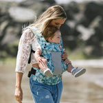 Tula - A woman carries a child on her chest wearing a Coast Paradise - Explore Baby Carrier. This is a side-on lifestyle shot in an outdoor environment. The baby is facing forward in the carrier.