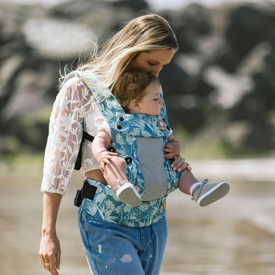 Tula - A woman carries a child on her chest wearing a Coast Paradise - Explore Baby Carrier. This is a side-on lifestyle shot in an outdoor environment. The baby is facing forward in the carrier.