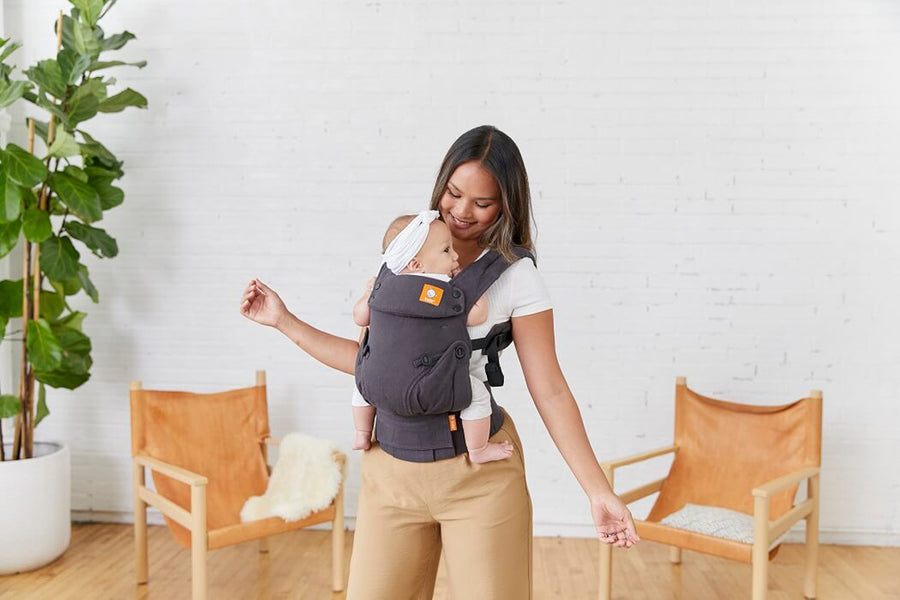 Woman stood in a white living room carrying a baby in a Tula explore grey baby carrier