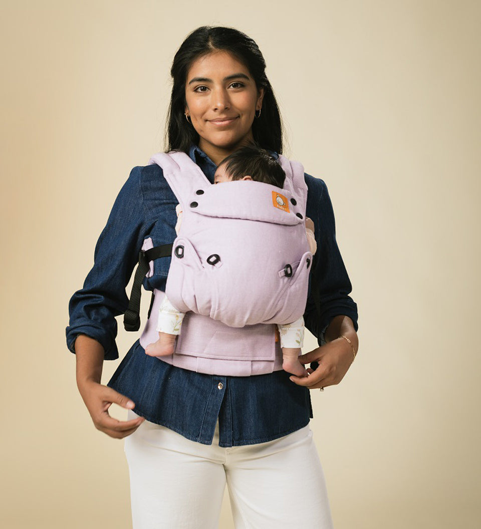 Woman stood on a beige background with a baby in a Tula explore herringbone baby carrier