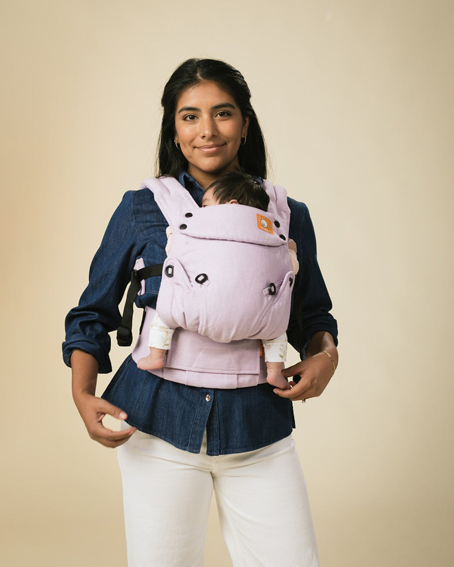 Woman stood on a beige background with a baby in a Tula explore herringbone baby carrier