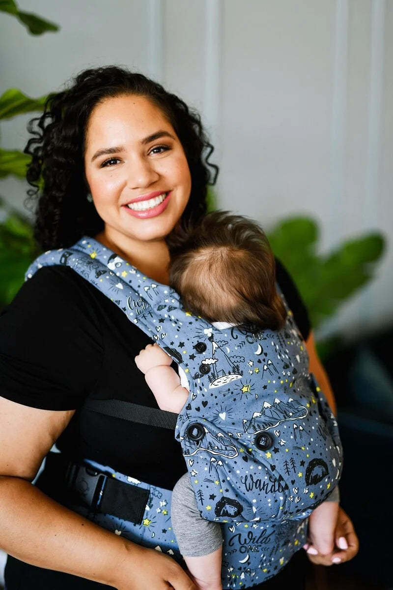 A person babywearing using the Tula Explore Baby Carrier featuring the Wander print. A blue background with black and white sketch drawings 