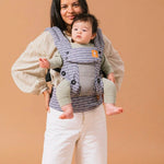 Woman stood in front of a brown background carrying a baby in a Tula explore coast beyond baby wearing carrier