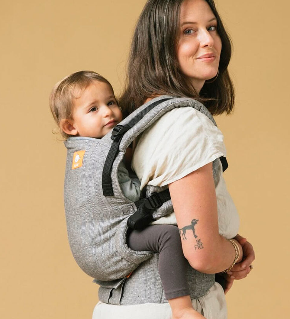 Woman stood on a yellow background carrying a baby in the Tula ash linen baby carrier