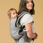 Woman stood on a yellow background carrying a baby in the Tula ash linen baby carrier