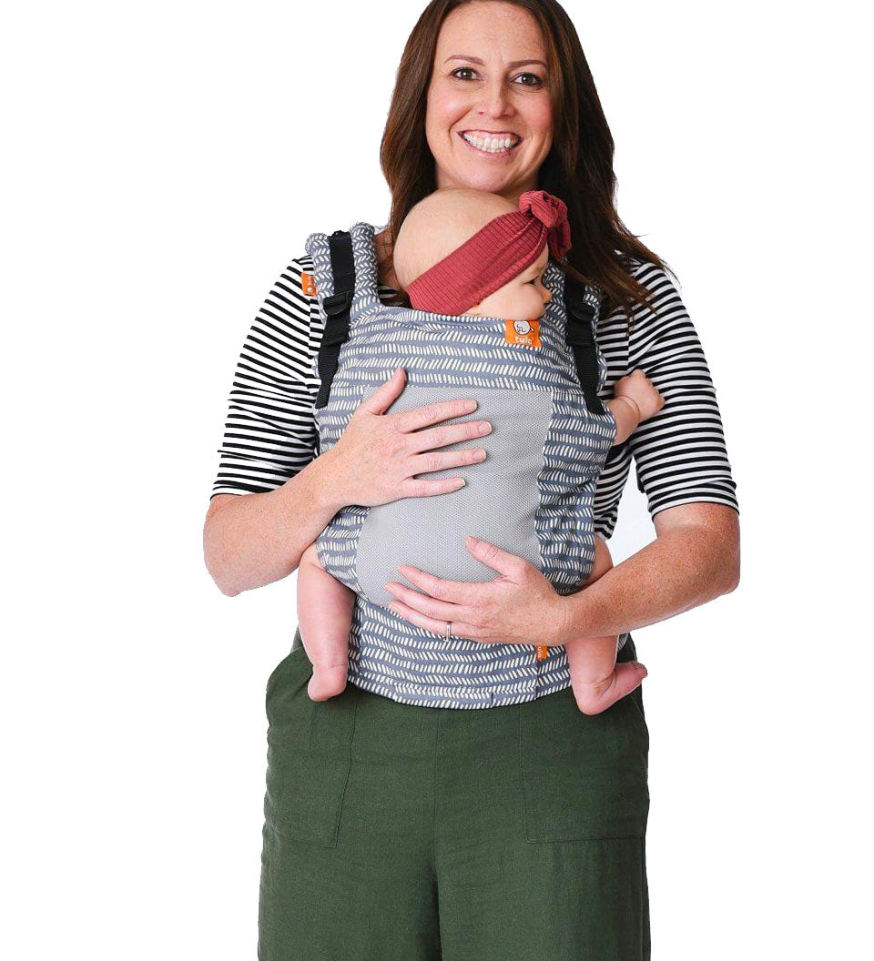 Woman stood on a white background carrying a baby in the Tula free to grow coast beyond baby carrier