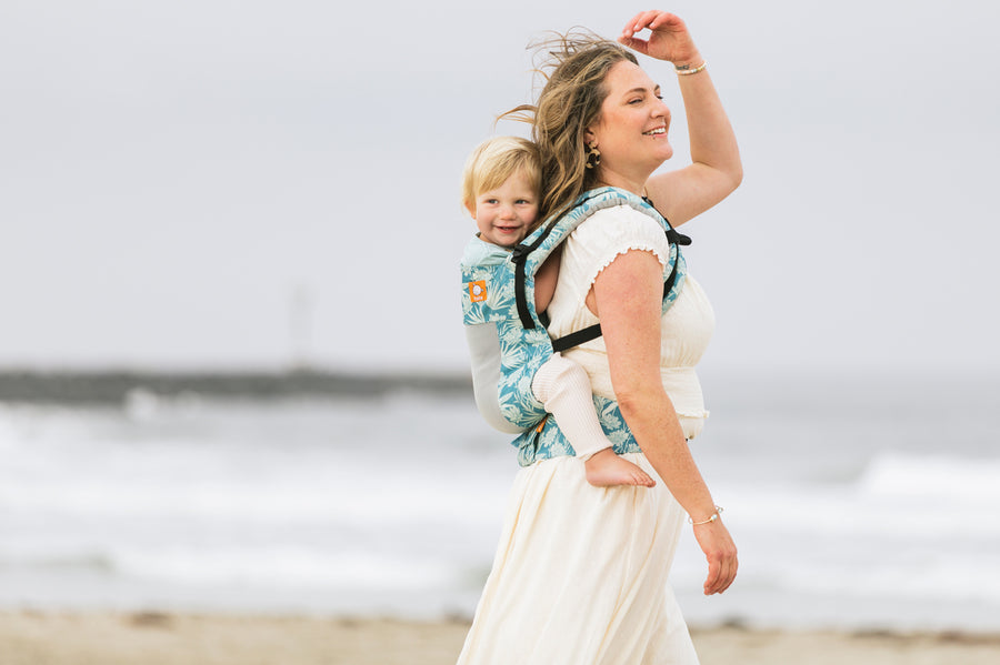 Woman walking along a beach wearing a baby on her back in the Tula soft structured FTG Coast Paradise baby carrier