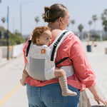 Woman walking on a beach wearing a baby on her back in the Tula FTG Coast Isle soft structured baby carrier