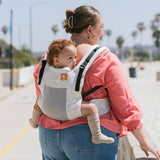 Woman walking on a beach wearing a baby on her back in the Tula FTG Coast Isle soft structured baby carrier