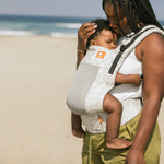 Close up of woman on a beach wearing the Tula beige Coast Isle FTG baby carrier