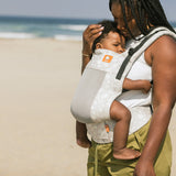 Close up of woman on a beach wearing the Tula beige Coast Isle FTG baby carrier