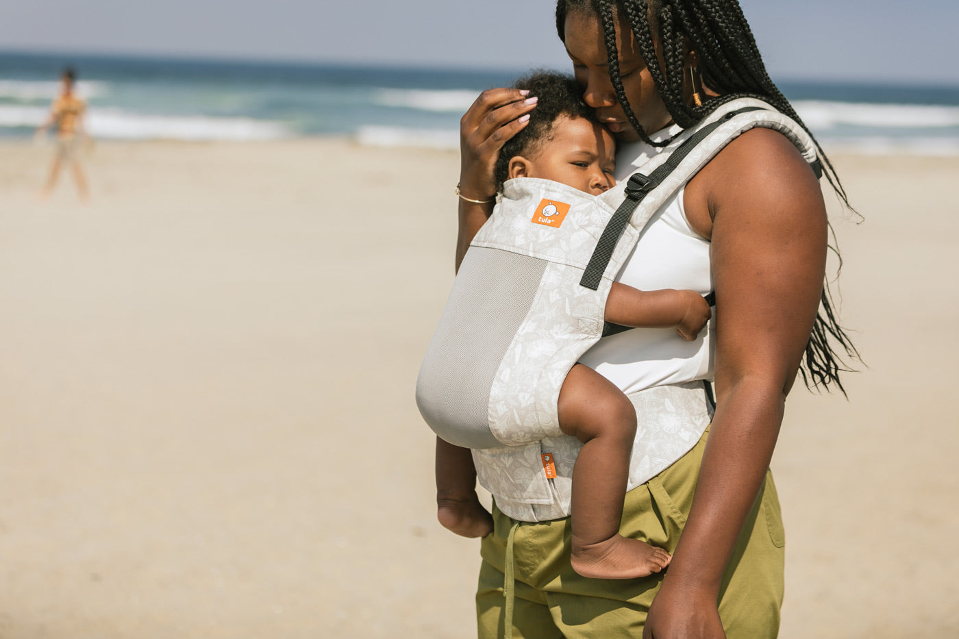 Close up of woman on a beach wearing the Tula beige Coast Isle FTG baby carrier