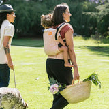 Woman walking in a grass field carrying a baby on her back in a Tula pink stardust soft baby carrier