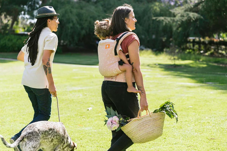 Woman walking in a grass field carrying a baby on her back in a Tula pink stardust soft baby carrier