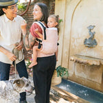 Woman stood in front of a water fountain carrying a child in a Tula pink stardust baby carrier