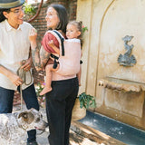 Woman stood in front of a water fountain carrying a child in a Tula pink stardust baby carrier