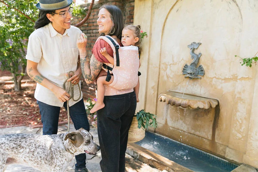 Woman stood in front of a water fountain carrying a child in a Tula pink stardust baby carrier