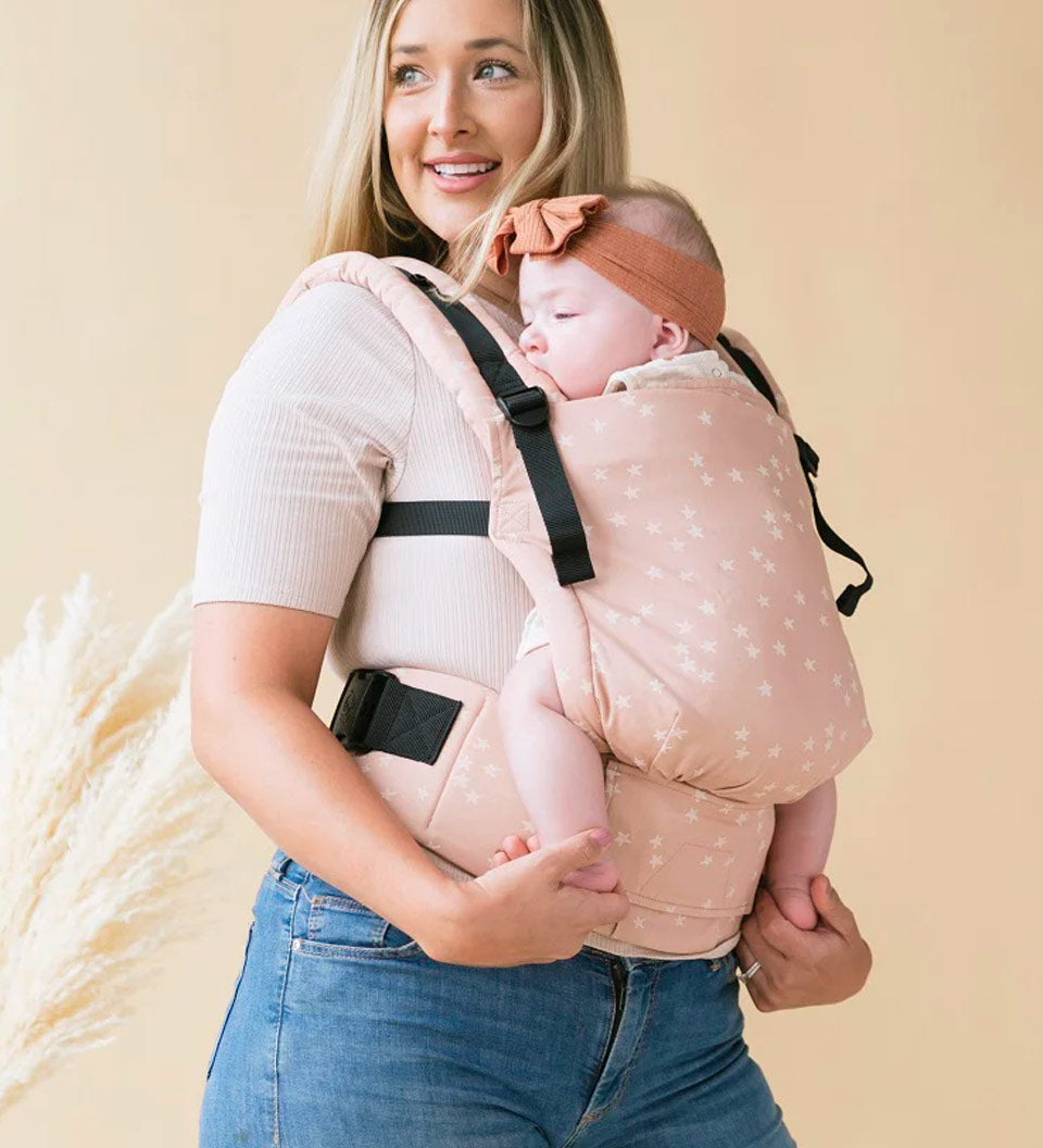 Woman carrying a baby in a Tula stardust FTG baby carrier on a beige background