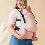 Woman carrying a baby in a Tula stardust FTG baby carrier on a beige background