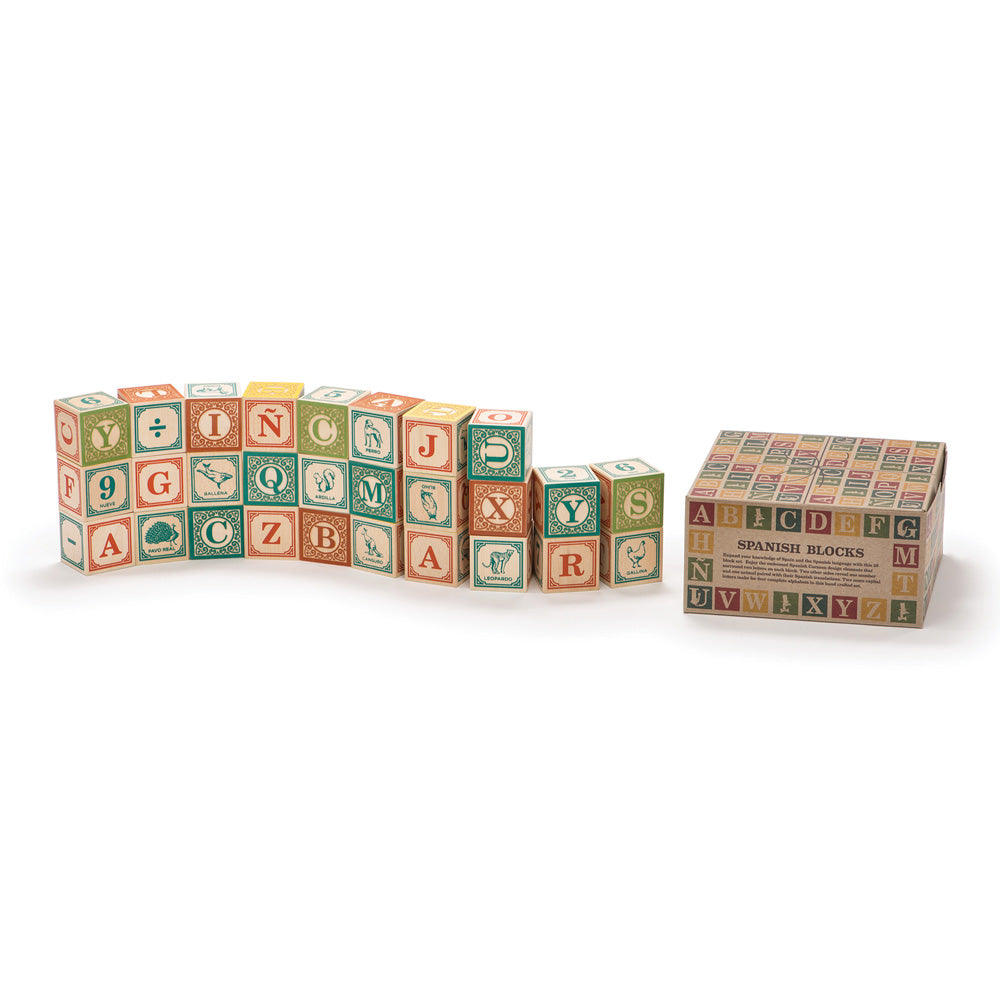 Uncle Goose wooden Spanish toy blocks stacked in a wavy pile next to their cardboard box on a white background