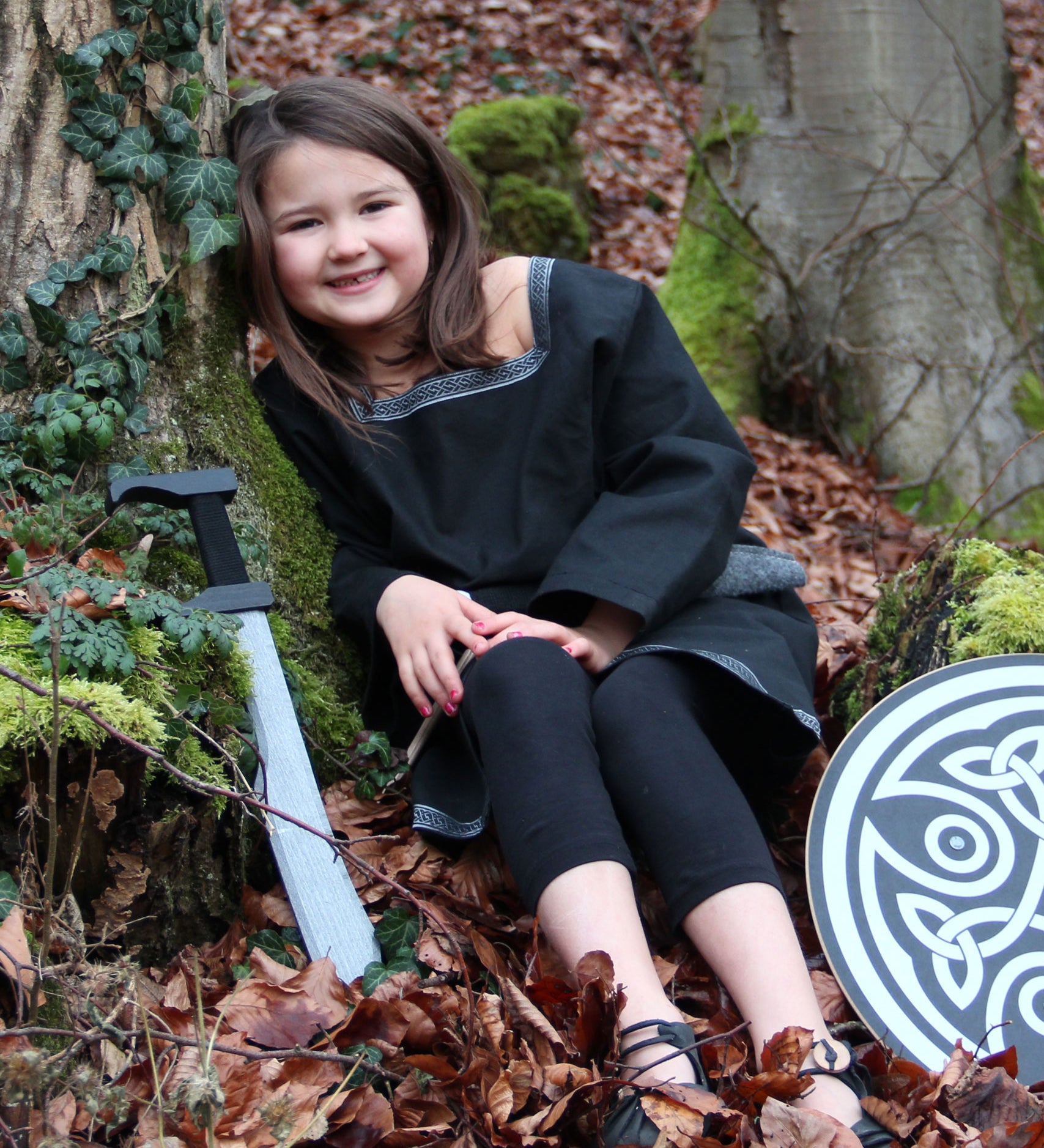 A child wearing a Vah Black Aragon child's Tunic, with a round shield and sword placed besides her on the floor. The child is sitting down on a forest.