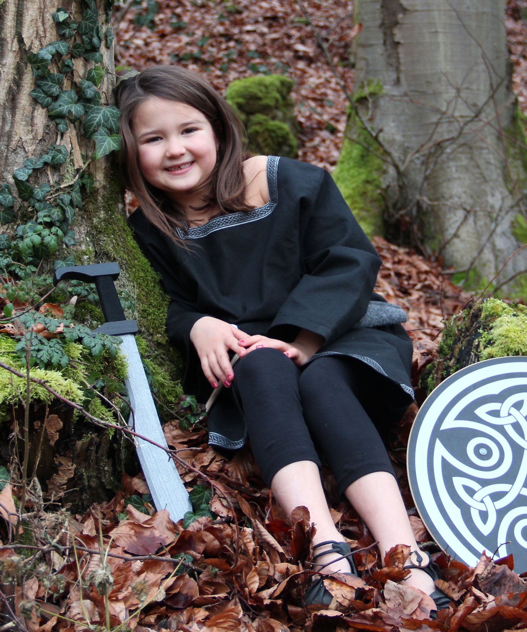 A child wearing a Vah Black Aragon child's Tunic, with a round shield and sword placed besides her on the floor. The child is sitting down on a forest.