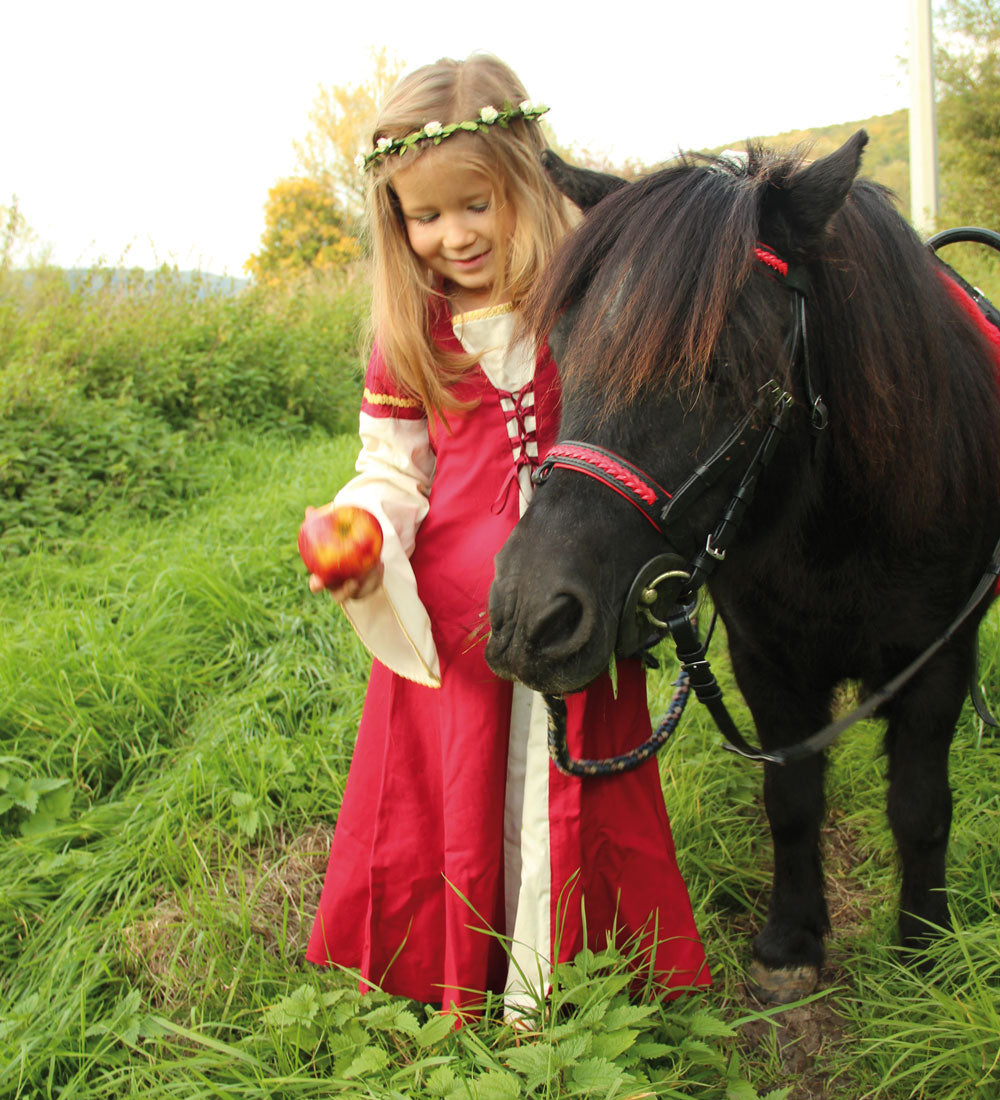 Child wearing the Vah Red Little Marian costume Dress with a cream coloured floral crown whilst feeding an apple to a pony