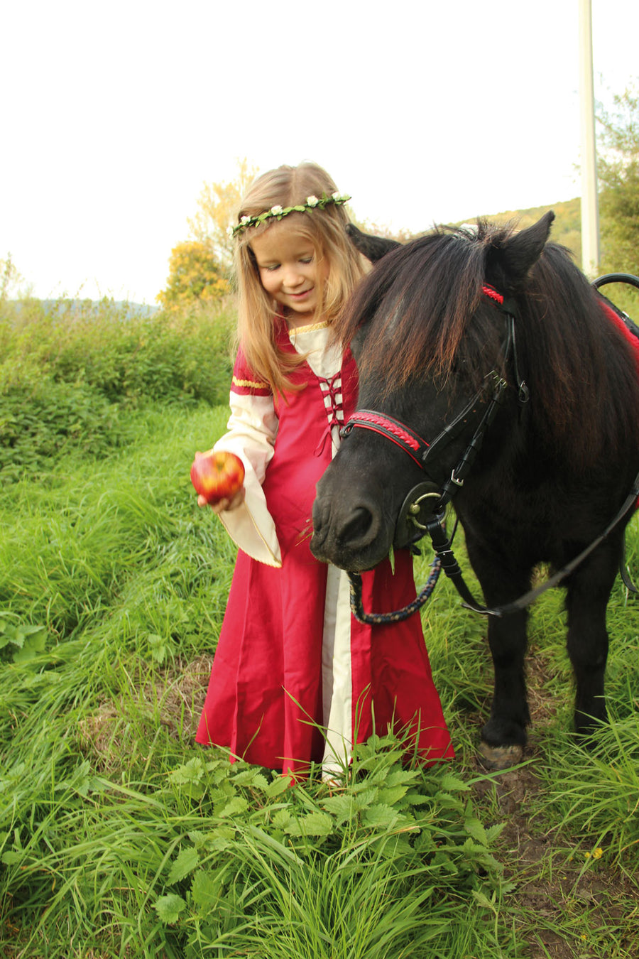 Child wearing the Vah Red Little Marian costume Dress with a cream coloured floral crown whilst feeding an apple to a pony